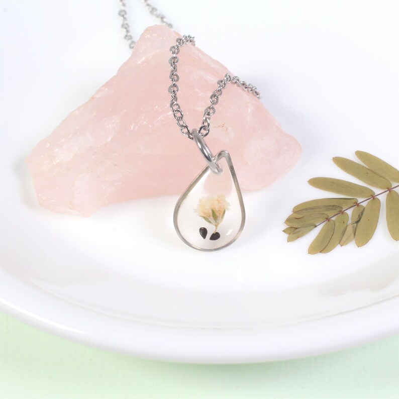 May include: A silver chain necklace with a teardrop-shaped pendant containing a pressed flower and seeds. The pendant is clear and the flower is white.