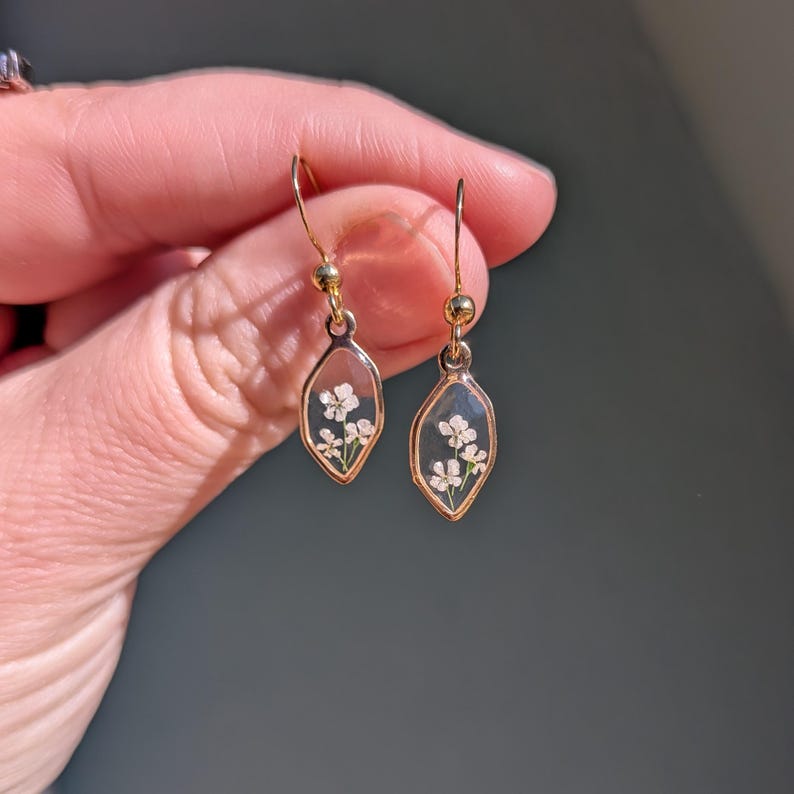 May include: Gold-colored dangle earrings with a clear, oval-shaped resin pendant. Each pendant contains small white flowers. The earrings are held by a person's hand against a blurred background.