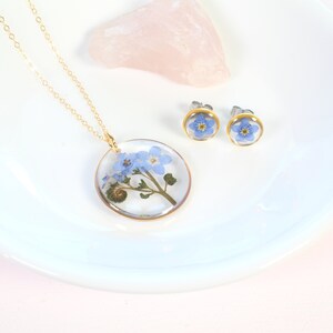 May include: A set of delicate jewelry featuring forget-me-not flowers pressed and preserved in clear resin. The set includes a necklace with a round pendant and a pair of stud earrings.