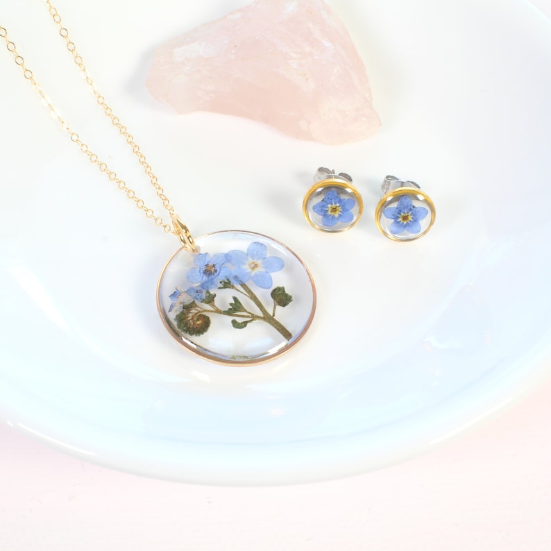 May include: A set of delicate jewelry featuring forget-me-not flowers pressed and preserved in clear resin. The set includes a necklace with a round pendant and a pair of stud earrings.