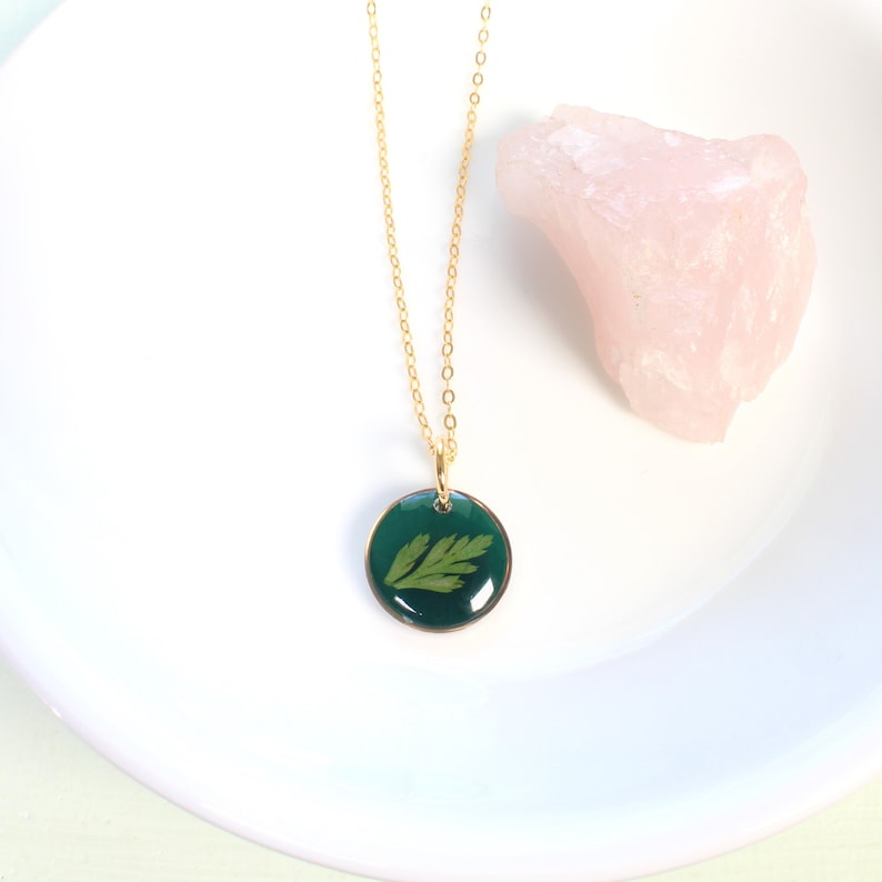 May include: A gold chain necklace with a round pendant featuring a pressed green leaf encased in clear resin.