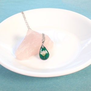 May include: A silver chain necklace with a teardrop-shaped pendant. The pendant is filled with green resin and a small white flower is embedded inside.