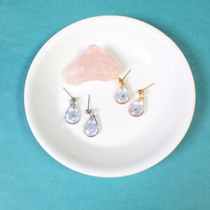 May include: Four pairs of earrings with a teardrop shape and a small blue flower pressed inside. The earrings are displayed on a white plate with a pink rock.