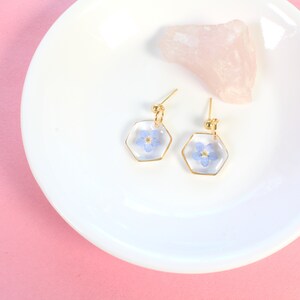 May include: A pair of gold hexagonal earrings with a delicate blue flower pressed inside each earring.