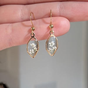 May include: A pair of gold-colored dangle earrings. Each earring features a clear, teardrop-shaped pendant with a delicate arrangement of small white flowers encased within. The earrings have a simple, elegant design.