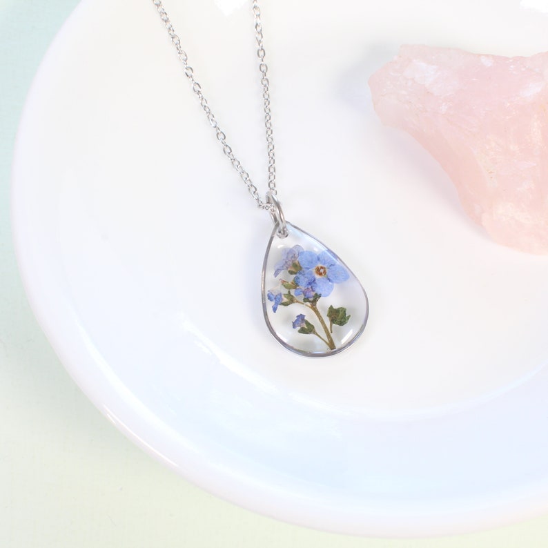 May include: A silver chain necklace with a teardrop-shaped pendant featuring a pressed forget-me-not flower encased in clear resin.