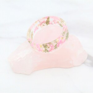 May include: A clear resin ring with pink and green pressed flowers embedded in the resin.