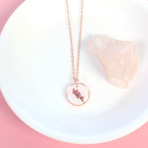 May include: A delicate rose gold necklace with a round pendant featuring a pressed flower encased in clear resin.