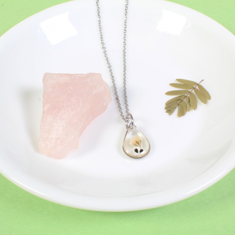 May include: A silver chain necklace with a teardrop-shaped pendant containing a pressed white flower. The pendant is on a white plate with a pink rock and a dried leaf.