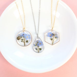 Peut inclure: Trois colliers délicats avec des fleurs de myosotis pressées à l'intérieur de pendentifs en résine transparente. Les pendentifs sont de forme ronde, en goutte d'eau et hexagonale. Les chaînes sont en or et en argent.