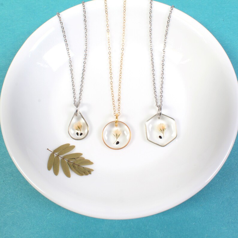May include: Three silver necklaces with different pendant shapes, each featuring a small white flower pressed inside clear resin. The pendants are a teardrop, a circle, and a hexagon.