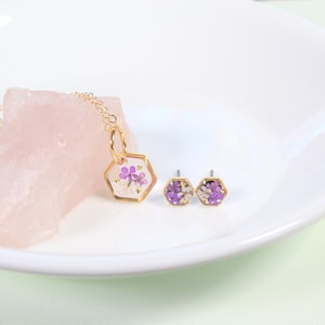 Op de afbeelding: Een gouden ketting met een zeshoekige hanger gevuld met geperste paarse en witte bloemen. Passende zeshoekige oorbel studs met hetzelfde bloemenontwerp.