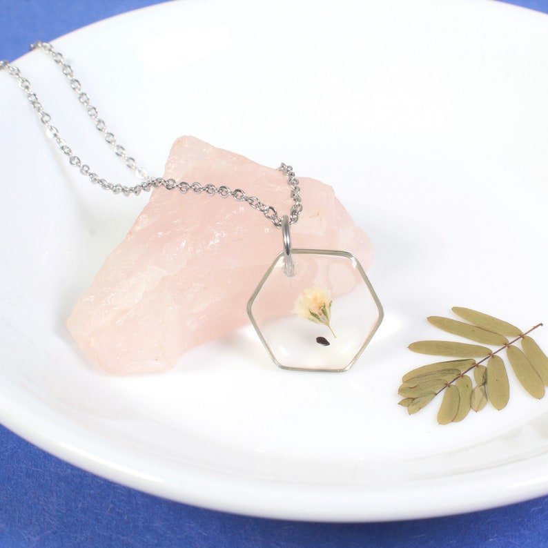 May include: A silver chain necklace with a hexagonal pendant containing a pressed flower. The pendant is clear and the flower is white with a pink center.