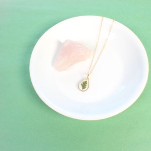 May include: A gold necklace with a teardrop-shaped pendant featuring a pressed green leaf. The necklace is displayed on a white plate with a pink quartz crystal.
