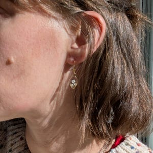 May include: A gold-colored earring with a small, clear, oval-shaped pendant. The pendant contains tiny white flowers. The earring is hanging from a person's ear.