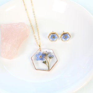 May include: A gold necklace and earrings set with a hexagonal pendant and earrings featuring pressed forget-me-not flowers encased in clear resin.