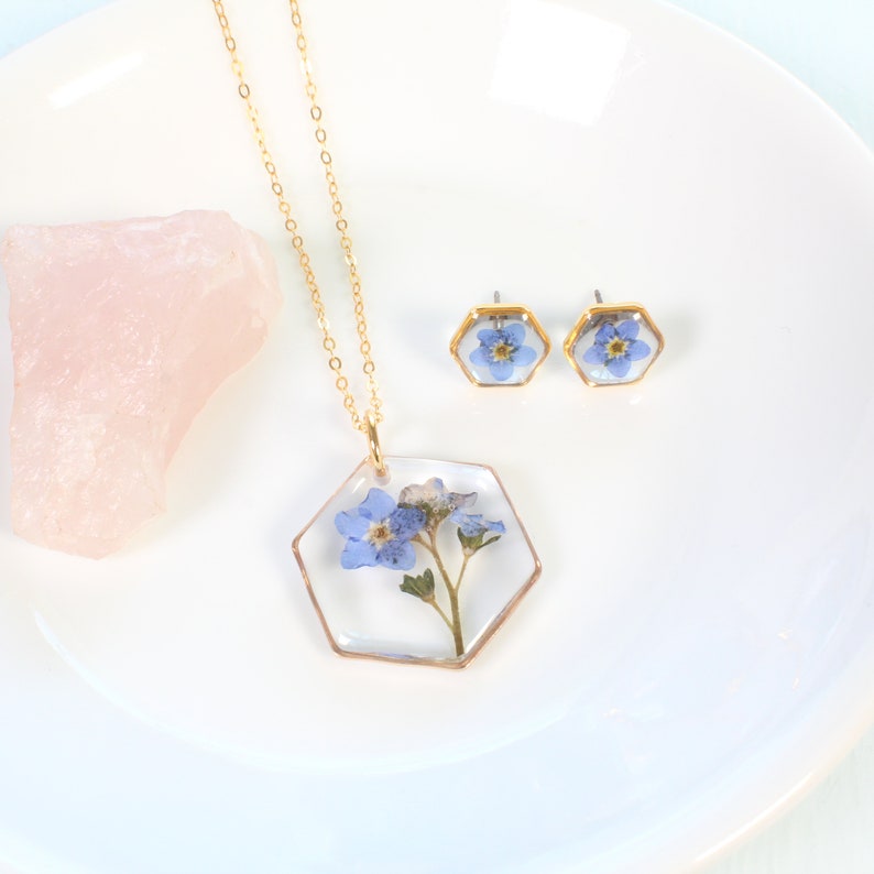 May include: A gold necklace and earrings set with a hexagonal pendant and earrings featuring pressed forget-me-not flowers encased in clear resin.