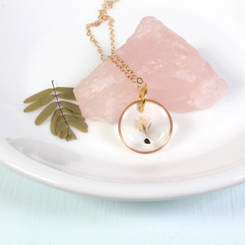 May include: A gold chain necklace with a round pendant featuring a small white flower pressed inside clear resin.