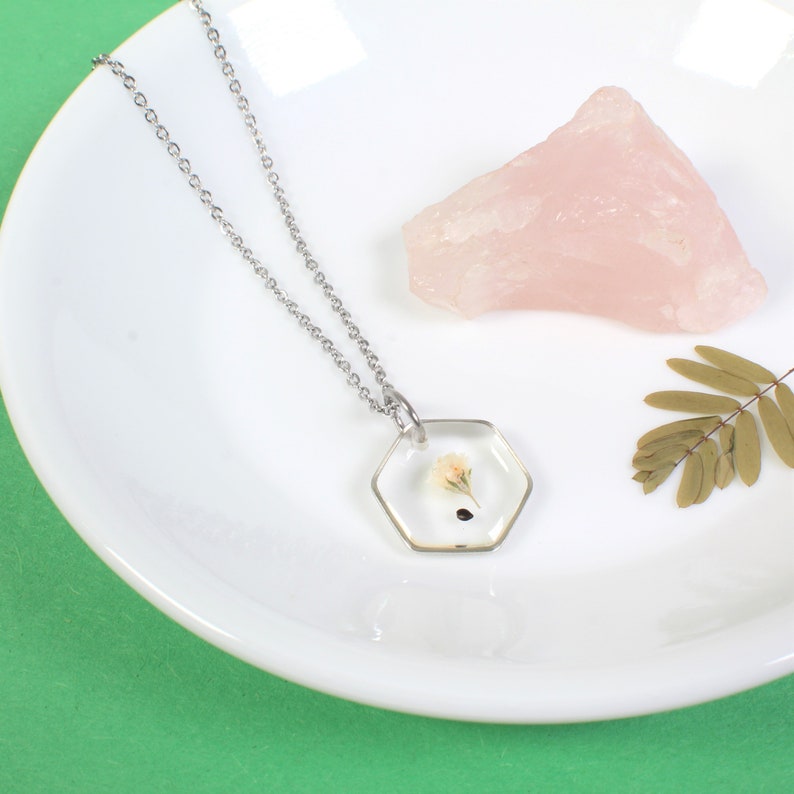 May include: A silver chain necklace with a hexagonal pendant containing a pressed white flower. The pendant is clear and the flower is visible inside.