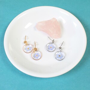 May include: Four pairs of earrings with small, round, clear resin settings containing pressed forget-me-not flowers. The earrings are displayed on a white plate with a pink crystal.