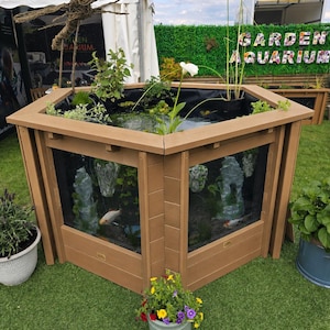 May include: A hexagonal garden aquarium with light brown wooden panels and clear glass viewing windows on each side. The top is open, revealing water, aquatic plants, and fish. A small grey pot with colorful flowers sits in the foreground.