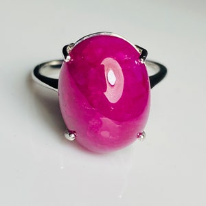 May include: A silver ring with a large, oval magenta gemstone. The polished gemstone reflects light. The ring's band is simple, polished silver. The gemstone's colour is a deep, rich magenta.