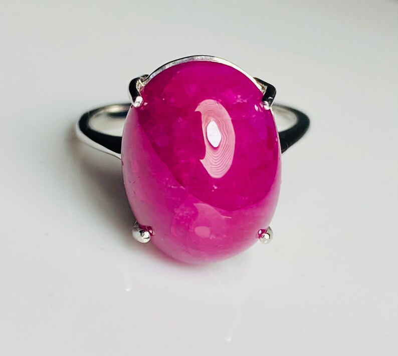 May include: A silver ring with a large, oval magenta gemstone. The polished gemstone reflects light. The ring's band is simple, polished silver. The gemstone's colour is a deep, rich magenta.