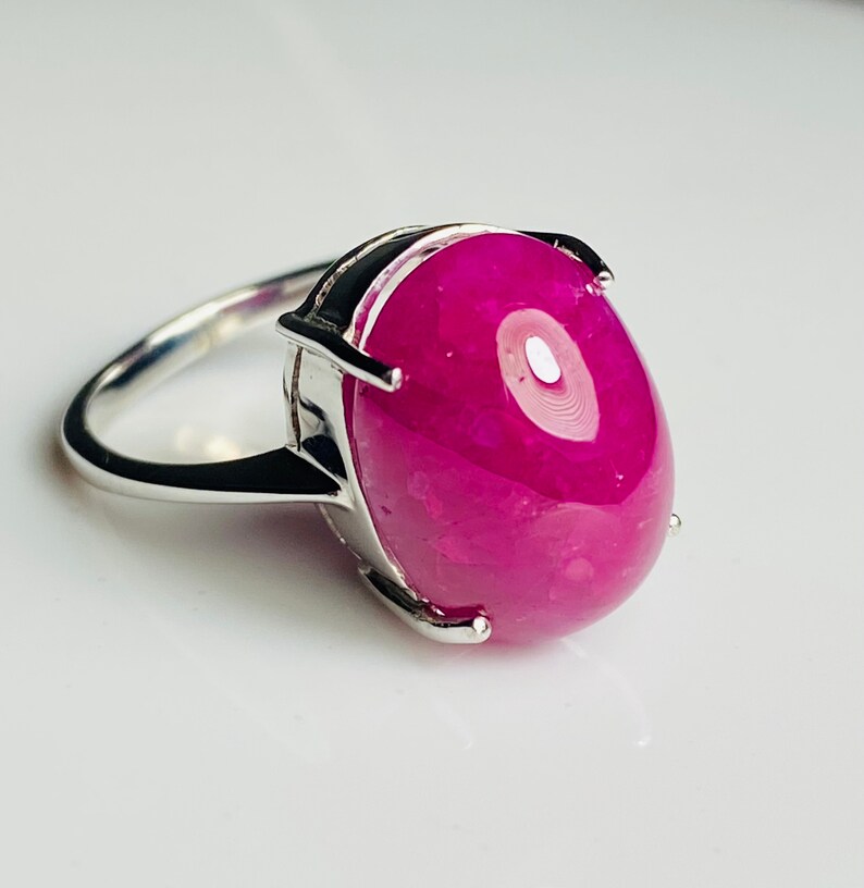 May include: A silver ring with a large, oval magenta gemstone. The gemstone is the focus, set in a silver band with a simple design. The ring is displayed on a white surface. The gemstone's colour is a deep, rich magenta.