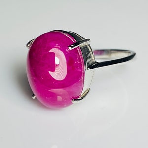 May include: A silver ring with a large, oval magenta gemstone. The gemstone is held in place by four prongs. The band is polished silver. The ring is shown against a white backdrop. The gemstone is approximately 2.5 cm in length.