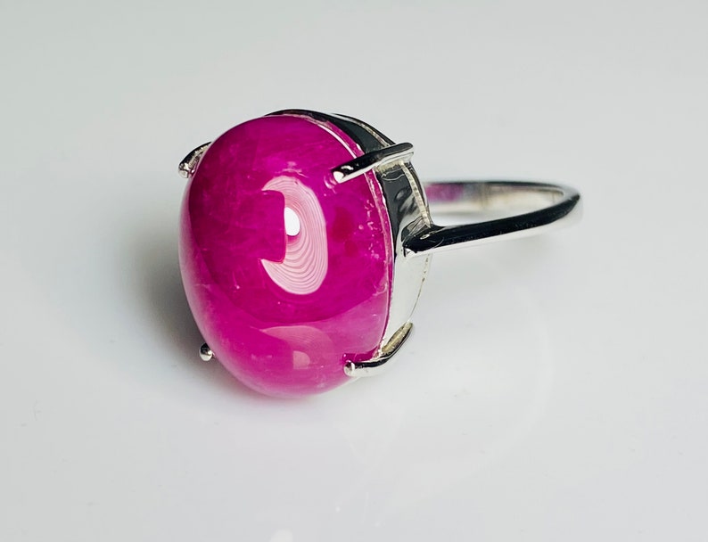 May include: A silver ring with a large, oval magenta gemstone. The gemstone is held in place by four prongs. The band is polished silver. The ring is shown against a white backdrop. The gemstone is approximately 2.5 cm in length.