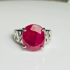 Puede incluir: Un anillo de plata con una gran gema de rubí de corte ovalado. El rubí es de color rojo intenso y está sujeto por cuatro garras. La banda del anillo tiene un diseño calado decorativo.