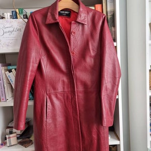 May include: A long, red leather jacket with a button closure and a collar. The jacket has a label that reads "Top Gun" on the inside.