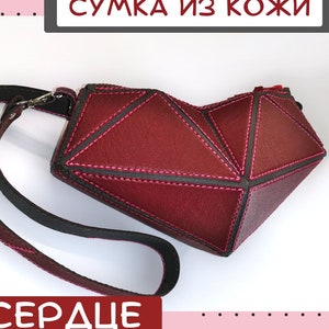 Könnte beinhalten: Eine rote Lederhandtasche in Herzform mit einem schwarzen Riemen. Die Tasche hat ein geometrisches Design mit rosa Nähten. Der Text "СУМКА ИЗ КОЖИ" ist in Schwarz auf rosa Hintergrund über der Tasche geschrieben. Der Text "СЕРДЦЕ" ist in Schwarz auf rosa Hintergrund unter der Tasche geschrieben.