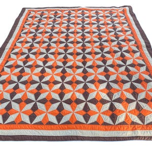 May include: A large, rectangular quilt with a geometric starburst pattern in shades of orange, brown, and gray. The quilt has a brown border and a gray stripe. The quilt is likely made of cotton or a similar fabric.