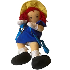 May include: A plush doll with red yarn hair, wearing a blue dress, white gloves, and black shoes. The doll has a yellow hat and a blue backpack with white and blue straps. The doll's legs are tan, and it has white socks with lace trim.