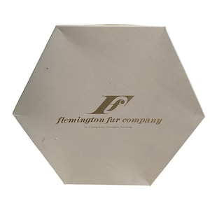 May include: A hexagonal off-white box featuring the gold-colored logo and text Flemington Fur Company, No. 8 Spring Street, Flemington, New Jersey printed on the center of the lid. The box has a smooth, matte finish and a clean, geometric shape.