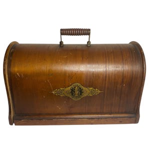 May include: A vintage Singer sewing machine case made of wood. The case has a curved top, a wooden handle, and a brass emblem with the Singer logo. The wood has a warm, brown tone with visible grain and some wear.
