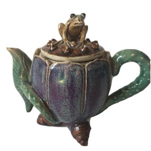 Pode incluir: Bule de cerâmica com uma figura de sapo na tampa. O bule é roxo e verde com um bico e uma alça em forma de folha. O sapo é marrom e o bule tem um design artístico único.