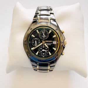 May include: A Seiko chronograph watch with a black dial, gold-tone accents, and a two-tone metal link band. The watch face features three sub-dials, a date window at the three o'clock position, and the text Seiko, Chronograph, and 50M.