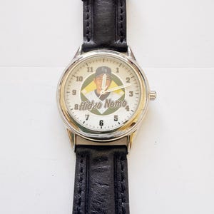 May include: A wristwatch with a silver-colored case and a black leather band. The watch face features a baseball player image with the name "Hideo Nomo" and the numbers 1 through 12. The watch has a classic design.