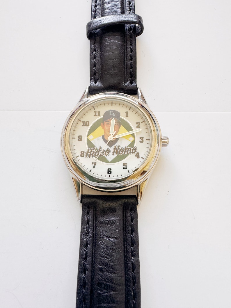 May include: A wristwatch with a silver-colored case and a black leather band. The watch face features a baseball player image with the name "Hideo Nomo" and the numbers 1 through 12. The watch has a classic design.
