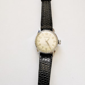 May include: A vintage wristwatch with a silver-colored case and a black leather band. The watch face is cream-colored with black numerals and hands. The watch reads "PAUL RAYNARD" and "17 JEWELS".