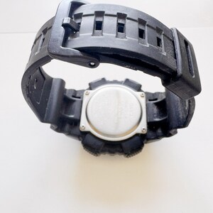 May include: Black digital watch with a black rubber strap. The watch face is silver with black buttons and a black bezel. The strap is open, showing the underside of the watch.