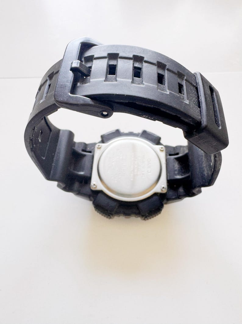 May include: Black digital watch with a black rubber strap. The watch face is silver with black buttons and a black bezel. The strap is open, showing the underside of the watch.
