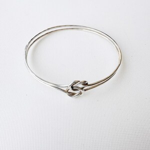 May include: A silver-toned bangle bracelet with a knot design at the center. The bracelet is made of two thin bands that meet at the knot. The background is a plain white surface.