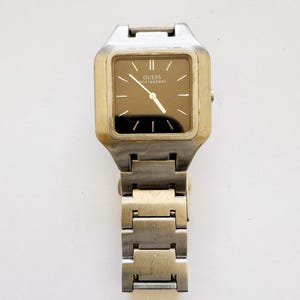May include: A vintage Guess wristwatch with a square, gold-toned face and a dark brown dial. The watch features silver-toned hands and hour markers. The band is made of silver-toned metal links. The watch face reads "GUESS microsteel".