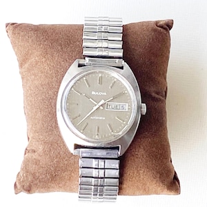 May include: A silver Bulova automatic wristwatch with a brown face and a silver metal band. The watch face has a day and date window and the text "Bulova Automatic" printed on it.