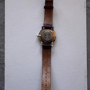 K&ouml;nnte beinhalten: Eine Armbanduhr mit braunem Lederarmband und silberner Schnalle. Das Zifferblatt der Uhr ist rund mit einem silbernen Geh&auml;use. Die R&uuml;ckseite der Uhr ist sichtbar. Das Armband ist etwa 1,8 cm breit.