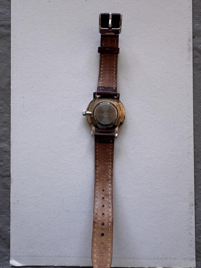 K&ouml;nnte beinhalten: Eine Armbanduhr mit braunem Lederarmband und silberner Schnalle. Das Zifferblatt der Uhr ist rund mit einem silbernen Geh&auml;use. Die R&uuml;ckseite der Uhr ist sichtbar. Das Armband ist etwa 1,8 cm breit.