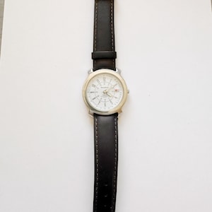 May include: A silver-toned wristwatch with a white face and black leather band. The watch has a round face with black numerals and hands. The black leather band is stitched and has a silver buckle.